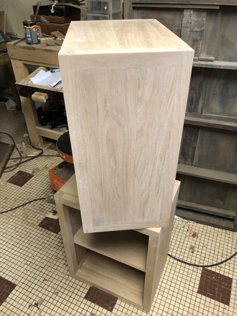 Mobilier en bois clair : une commode en bois brut surmontée d'un cube. Atelier d'ébénisterie en arrière-plan.